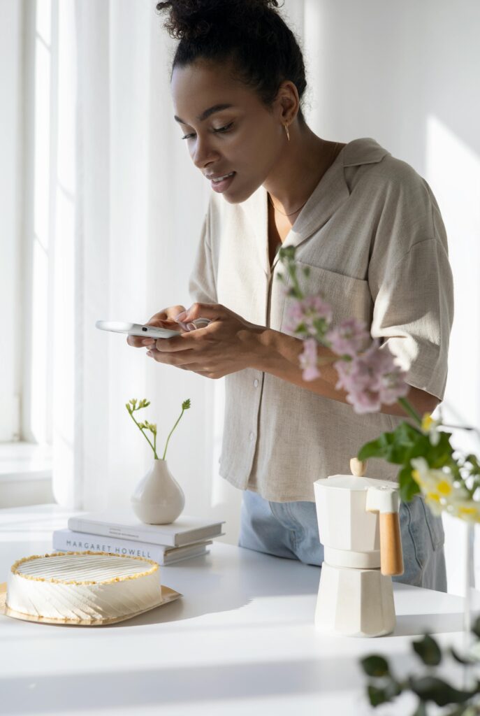 Woman capturing a stylish coffee setup in a sunlit kitchen with flowers and cake.